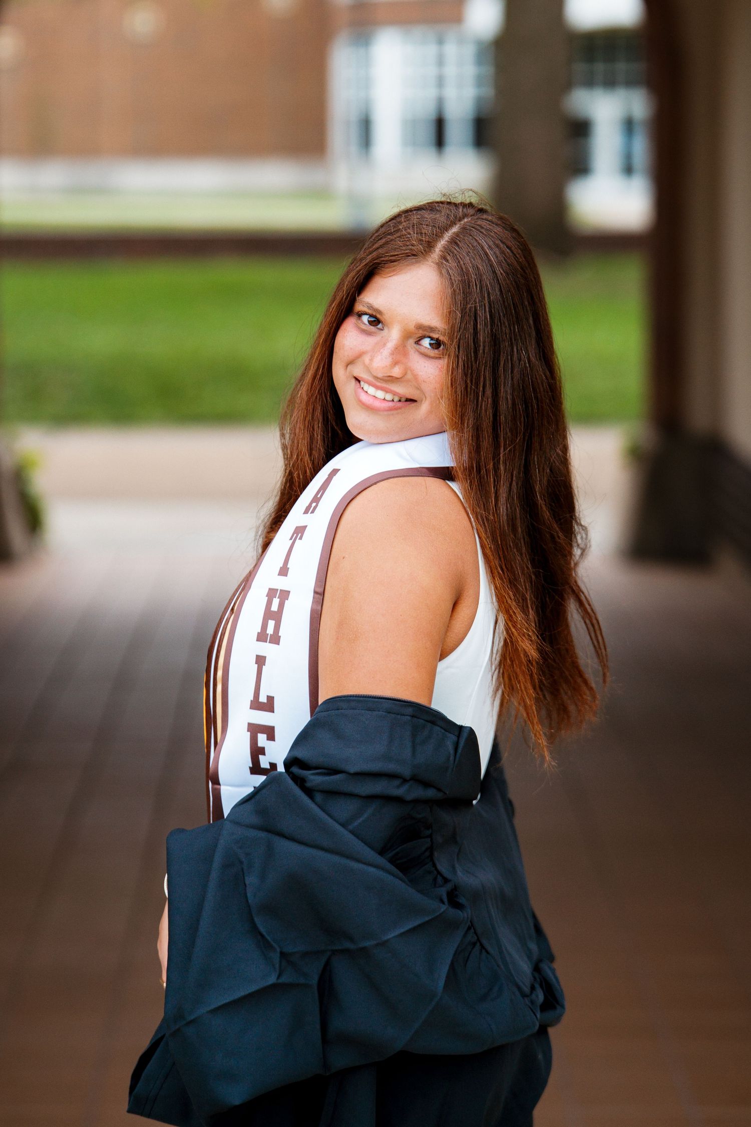 A smiling graduate in a white top and Athlete stole looks over her shoulder while holding a black gown under a covered walkway.