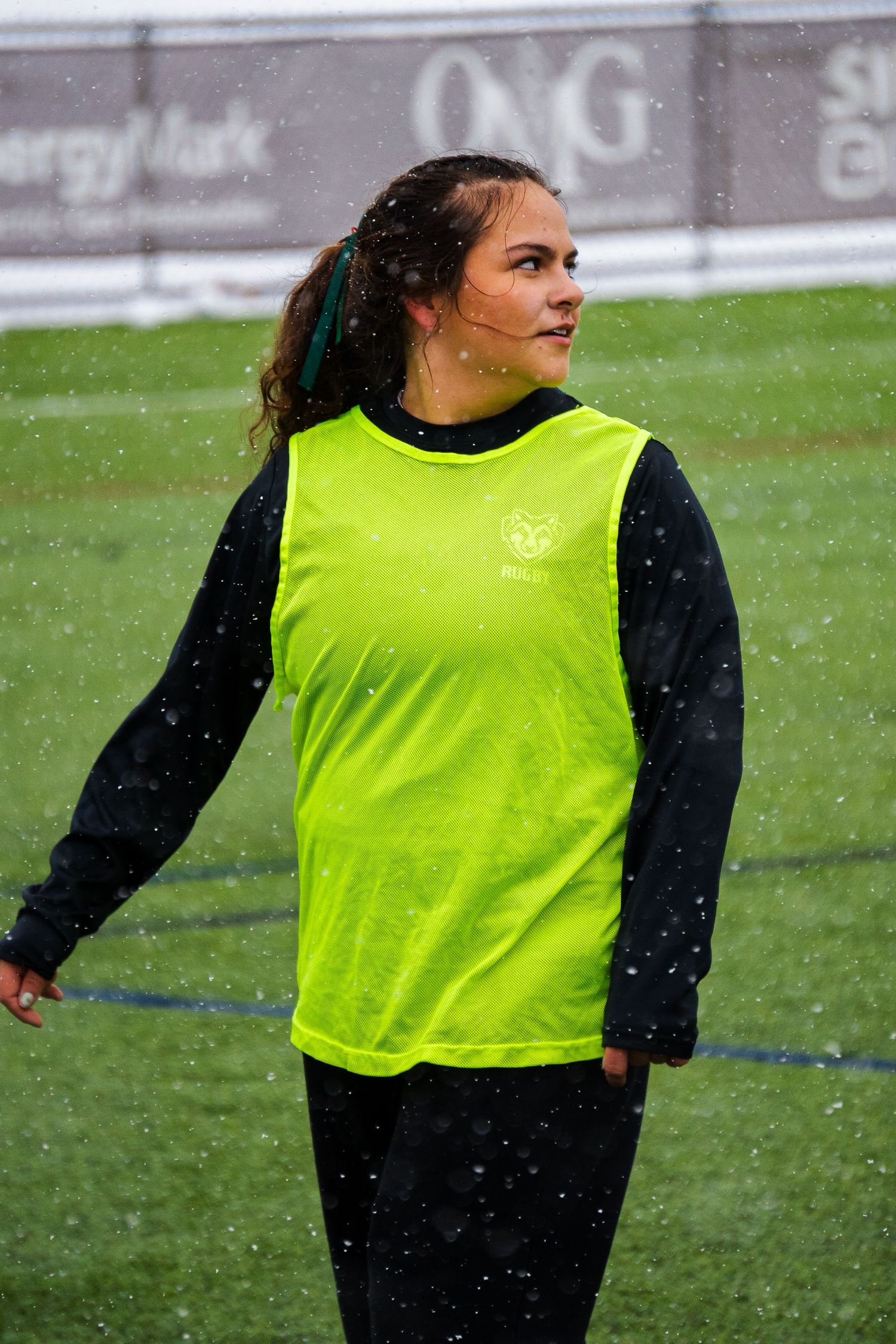 A women's rugby player in a neon green training bib looks off to the side as snow falls on the turf field during practice.