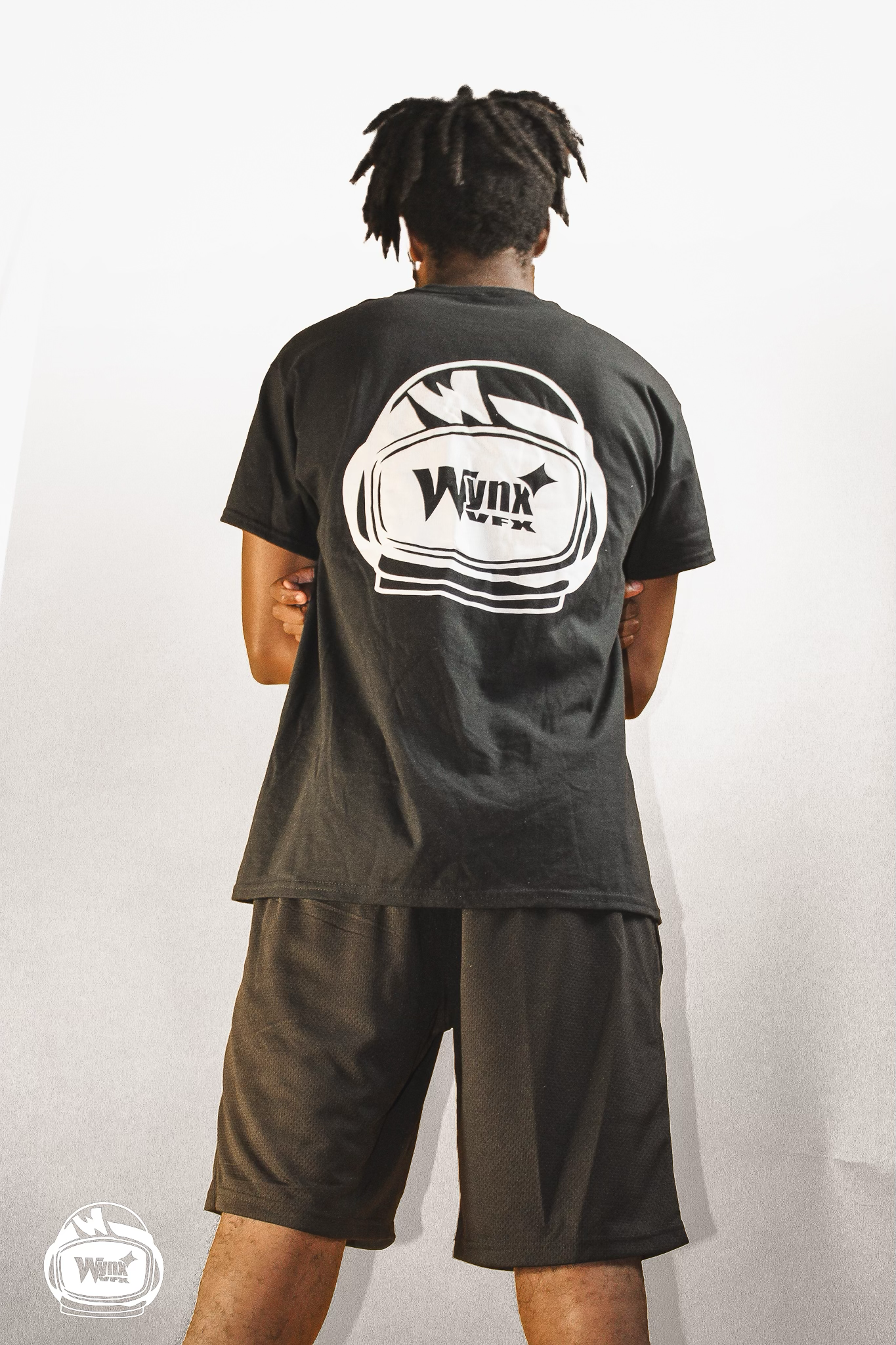 Man seen from behind wears a black t-shirt with a white Wynx helmet logo and black shorts against a plain white backdrop.