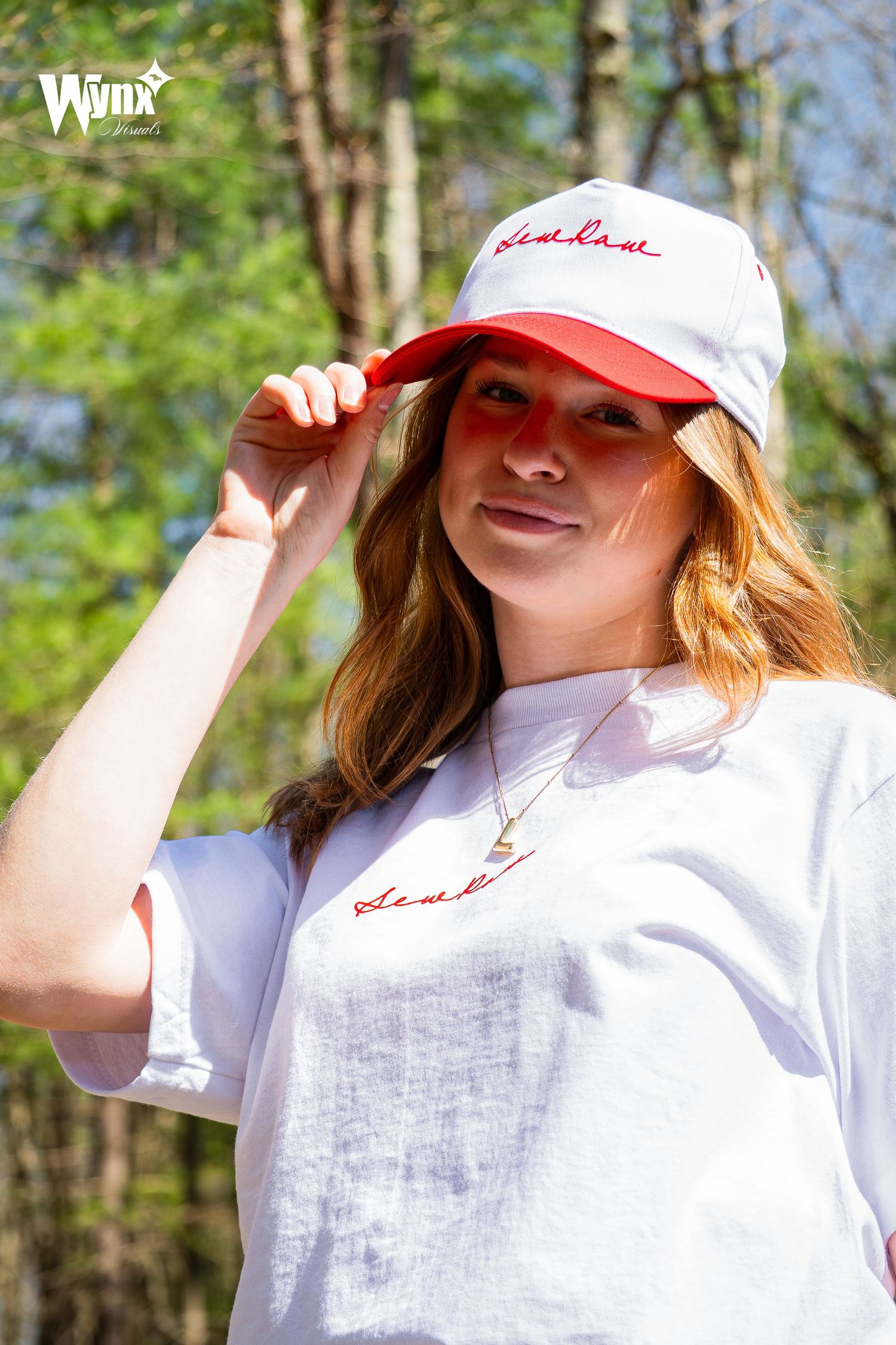 Smiling woman tipping the brim of a white and red Sewlaw cap, wearing a white tee with red script lettering among green trees.