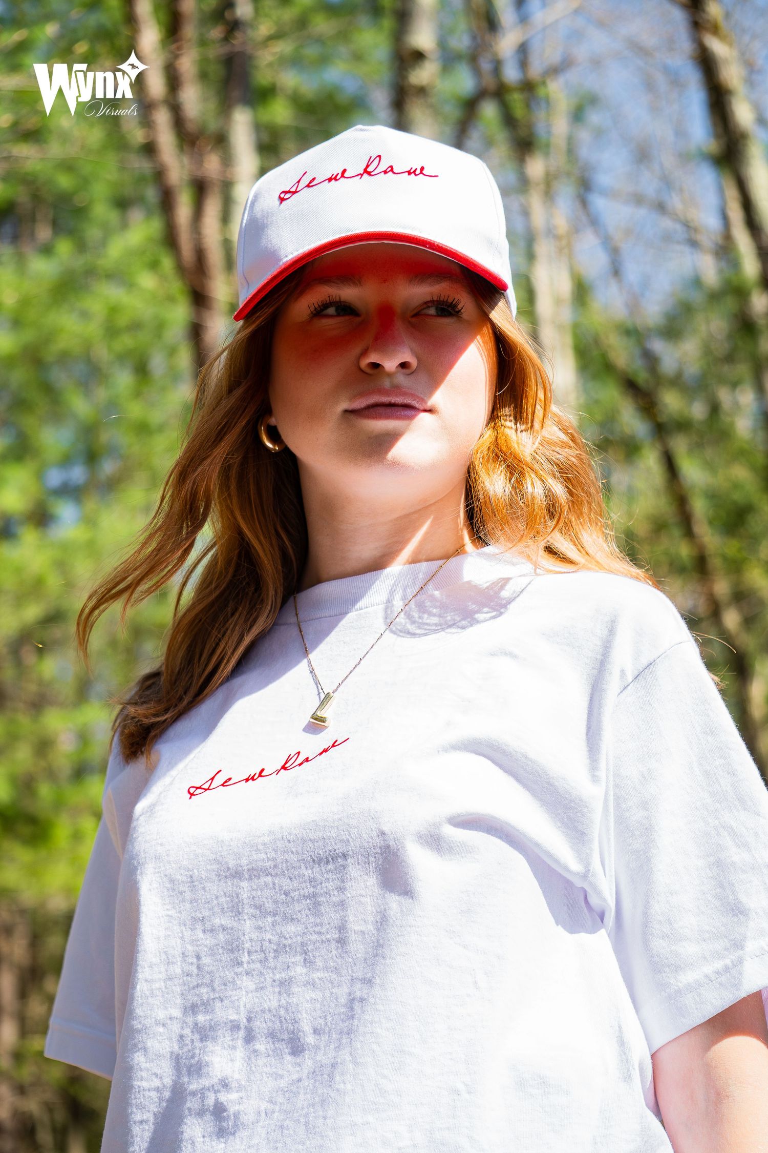 Woman with long auburn hair wearing a white and red Sewlaw cap and white script tee, standing in a sunlit forest.
