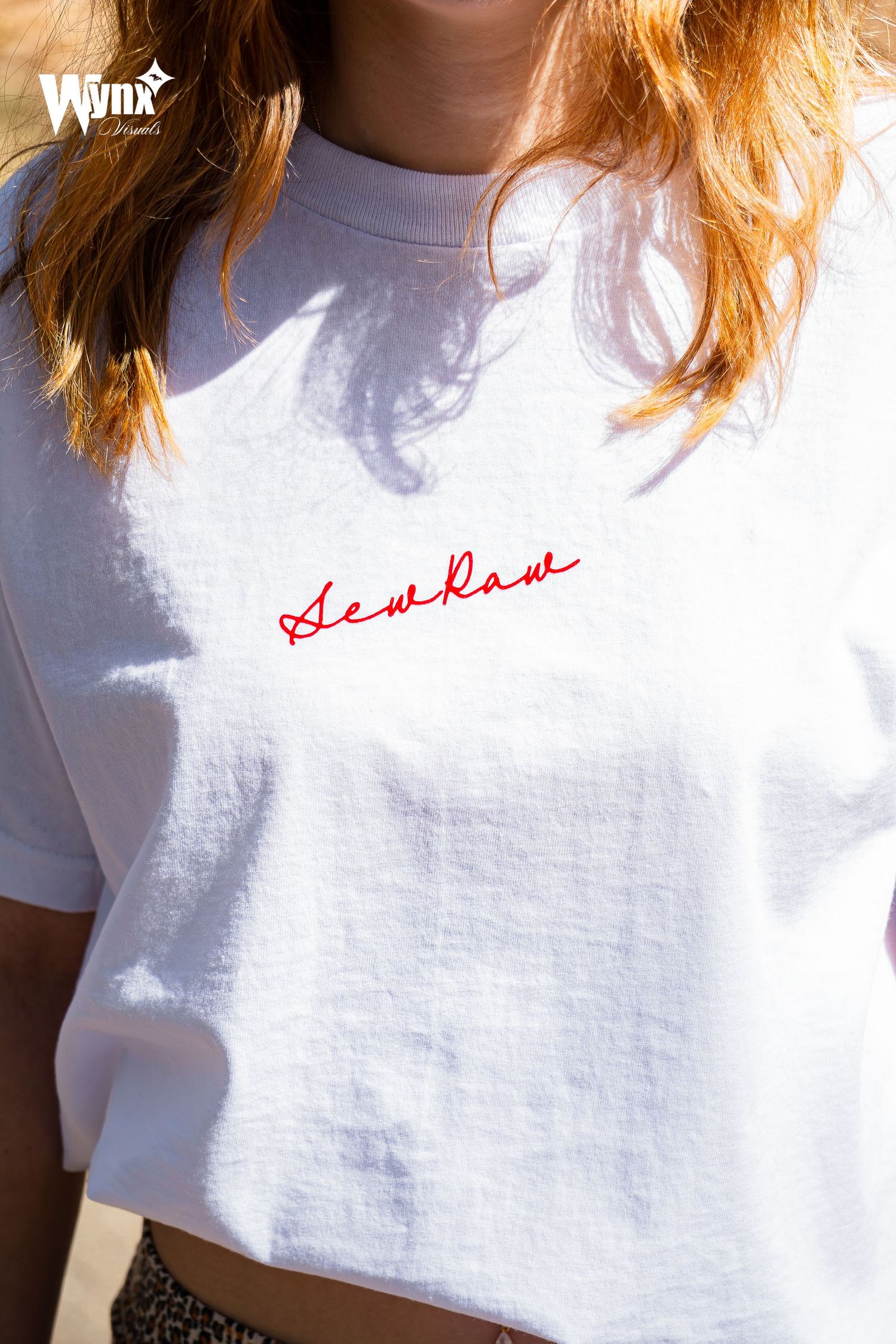 Close-up of a person in a white oversized t-shirt with red cursive script lettering on the chest, with sunlit auburn hair falling forward.