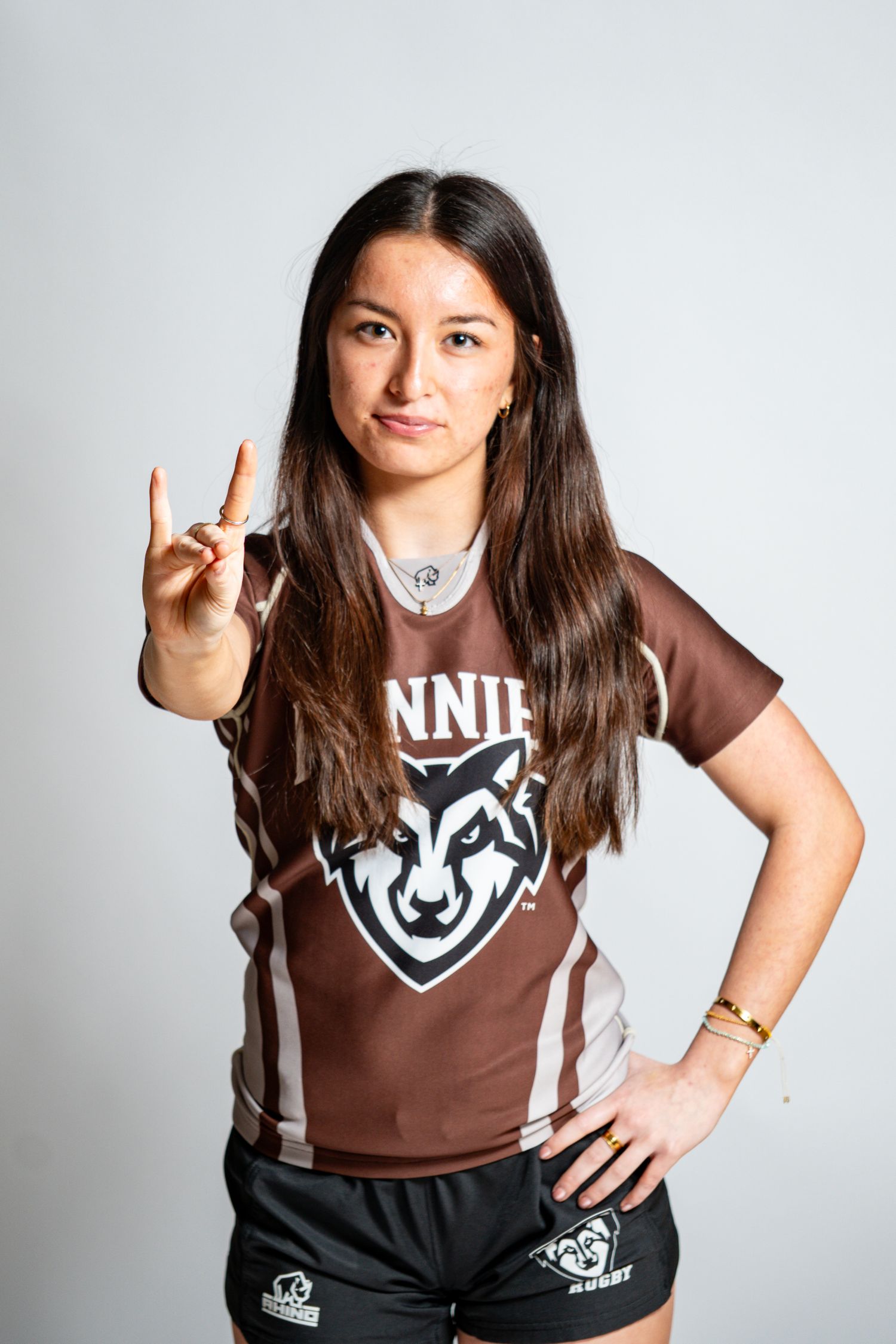 Woman in a brown rugby jersey with a wolf logo poses with a hand sign and her other hand on her hip against a white backdrop.
