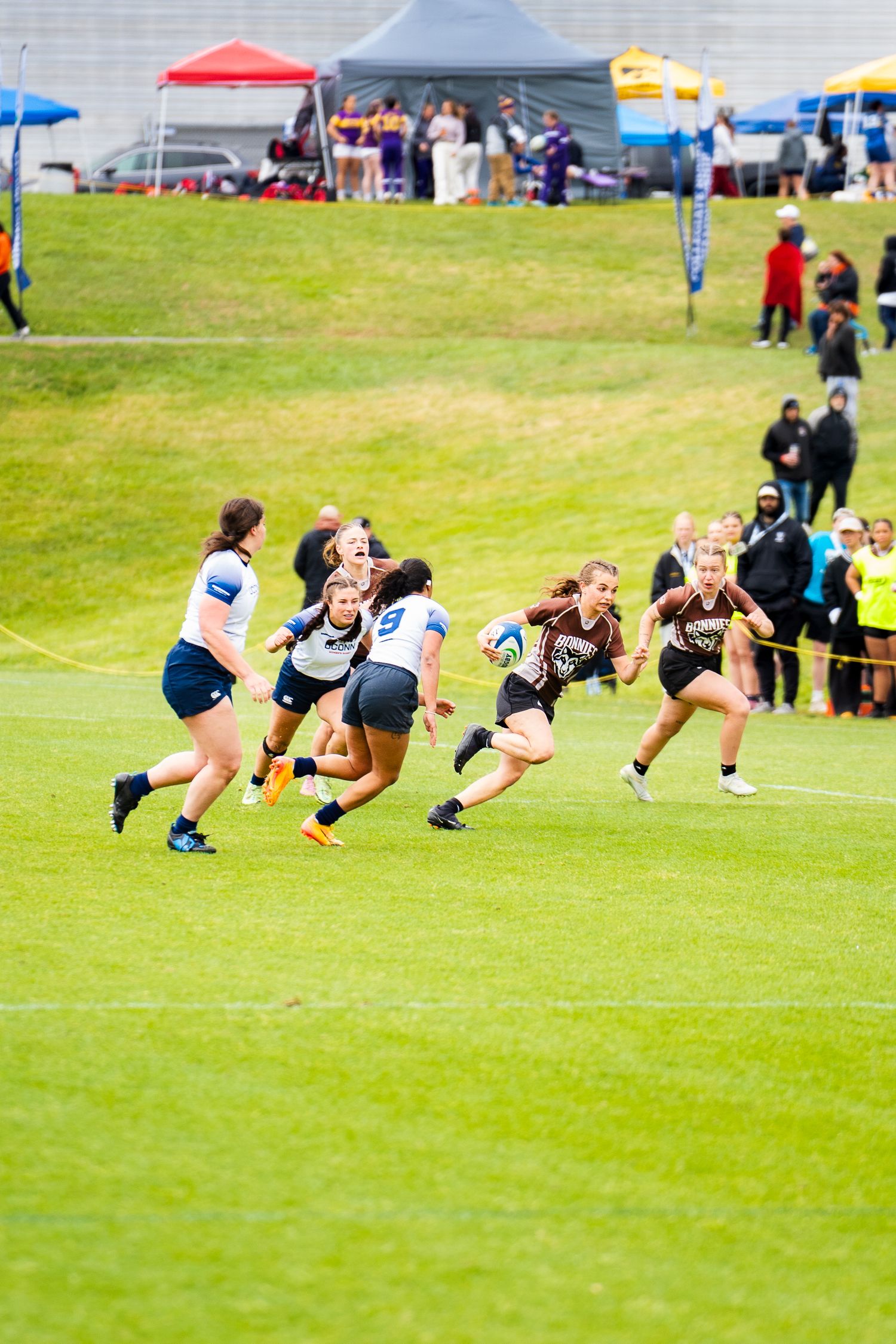A women's rugby player in a brown jersey runs with the ball as defenders in white chase her while fans watch from a grassy hillside.