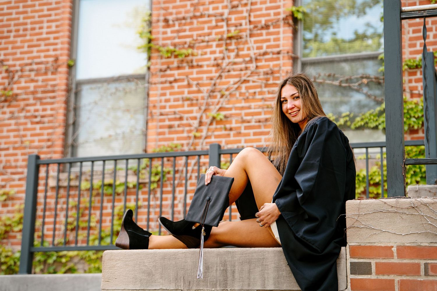 Graduate in a black gown and boots sits on brick steps beside a wrought-iron railing, holding her graduation cap.