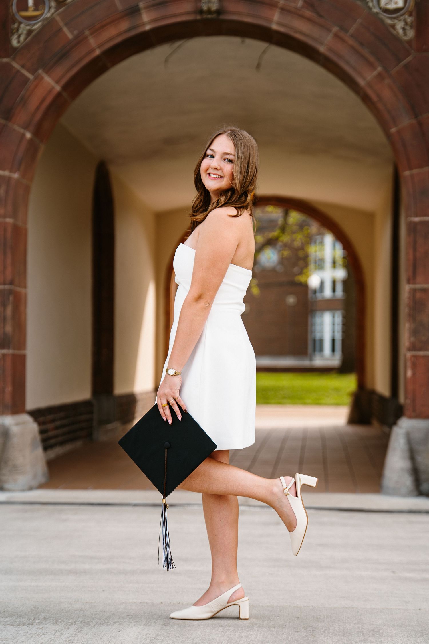 Smiling graduate in a white strapless dress and heels holds her black cap and kicks up one foot under a brick archway.