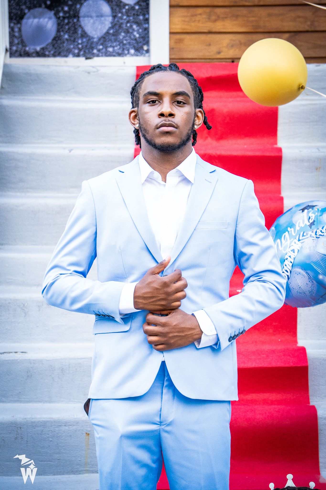 Young man in a light blue suit and white shirt stands on a red carpet beside white steps, with yellow and blue balloons nearby.