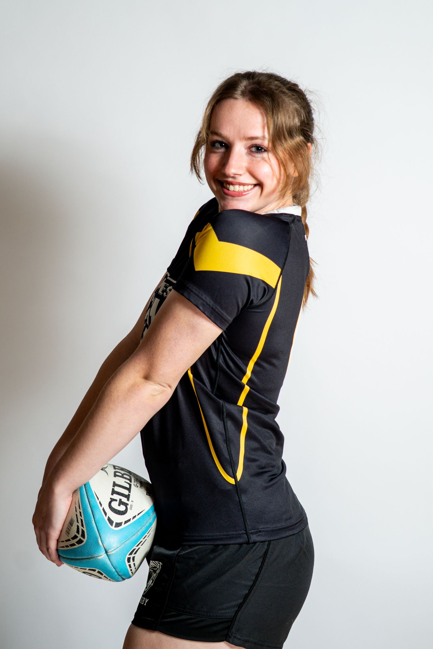 Smiling rugby player in a black and gold jersey holds a blue and white Gilbert rugby ball against her hip in a studio portrait.