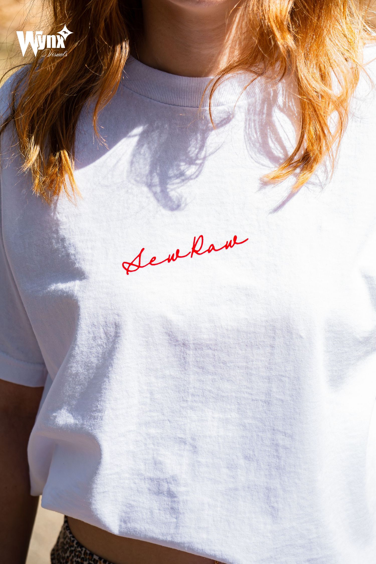 Close-up of a woman in a white oversized t-shirt with red cursive embroidered lettering on the chest in warm sunlight.