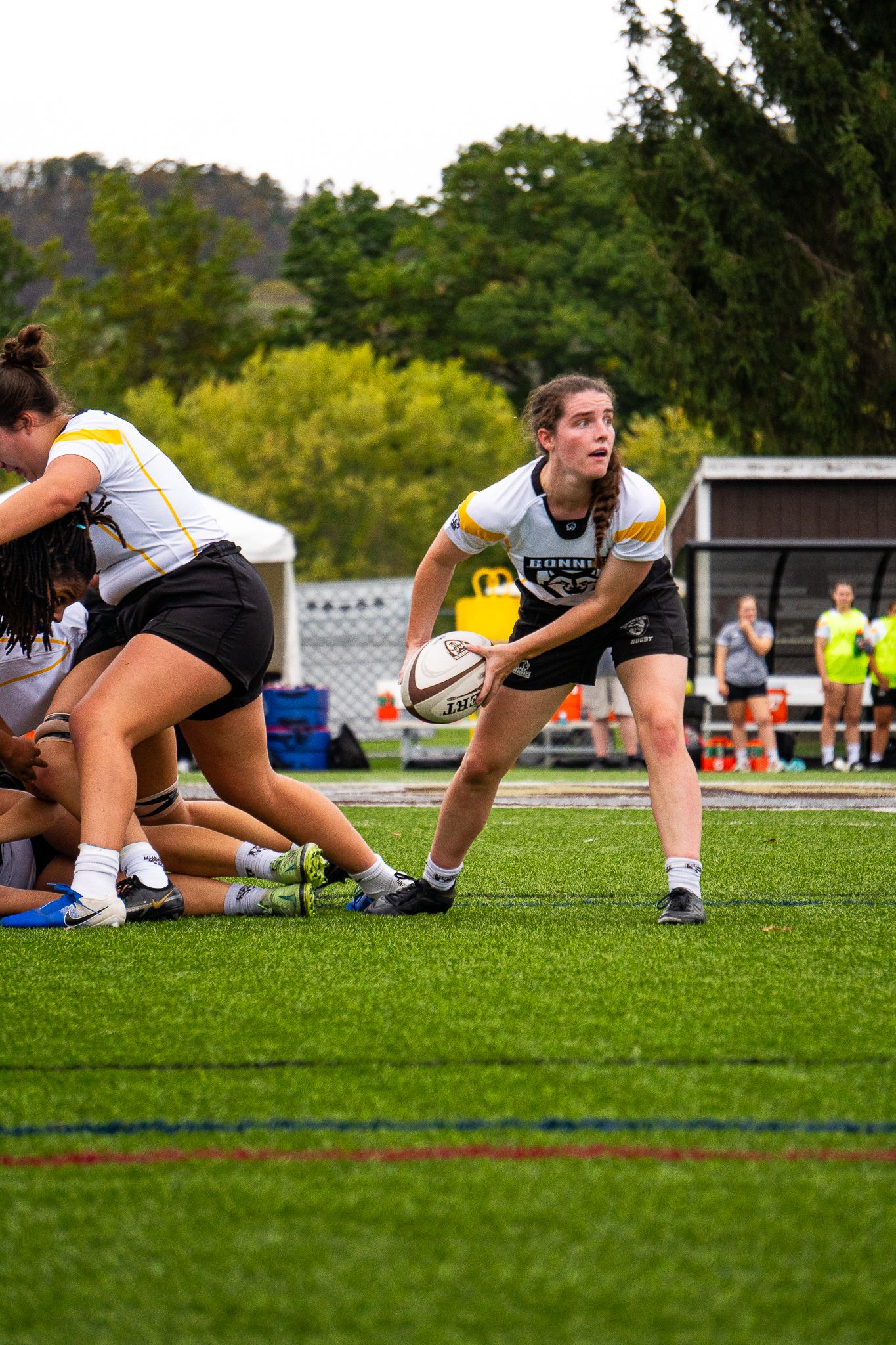 A Bonnies women's rugby scrumhalf in a white and gold jersey prepares to pass the ball from the ruck on a turf field.