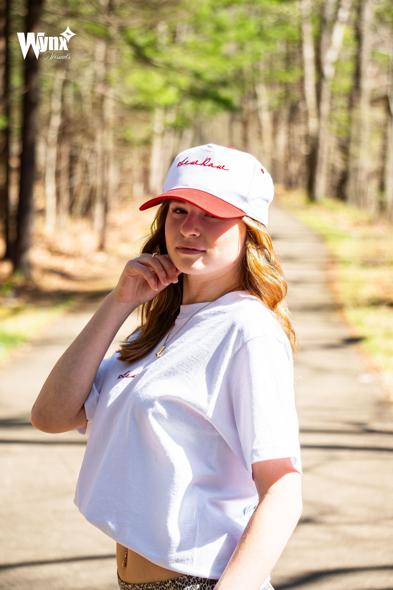 Woman in a white and red Sewlaw cap and cropped white tee posing with her hand near her face on a sunny forest road.