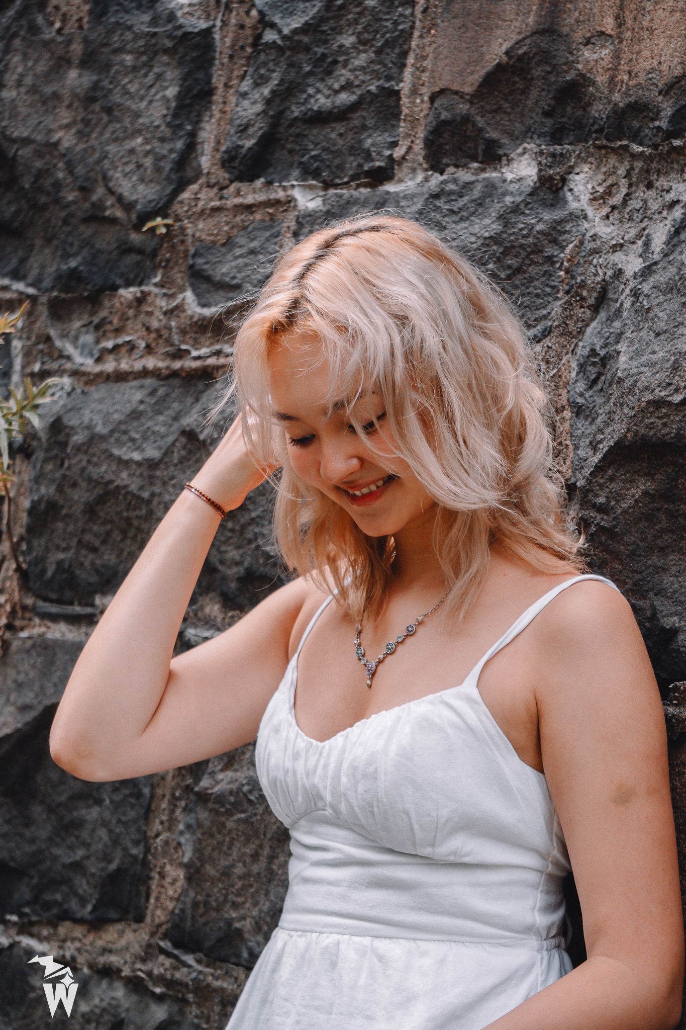 Young woman with blonde wavy hair in a white sundress smiling and touching her hair against a dark stone wall.