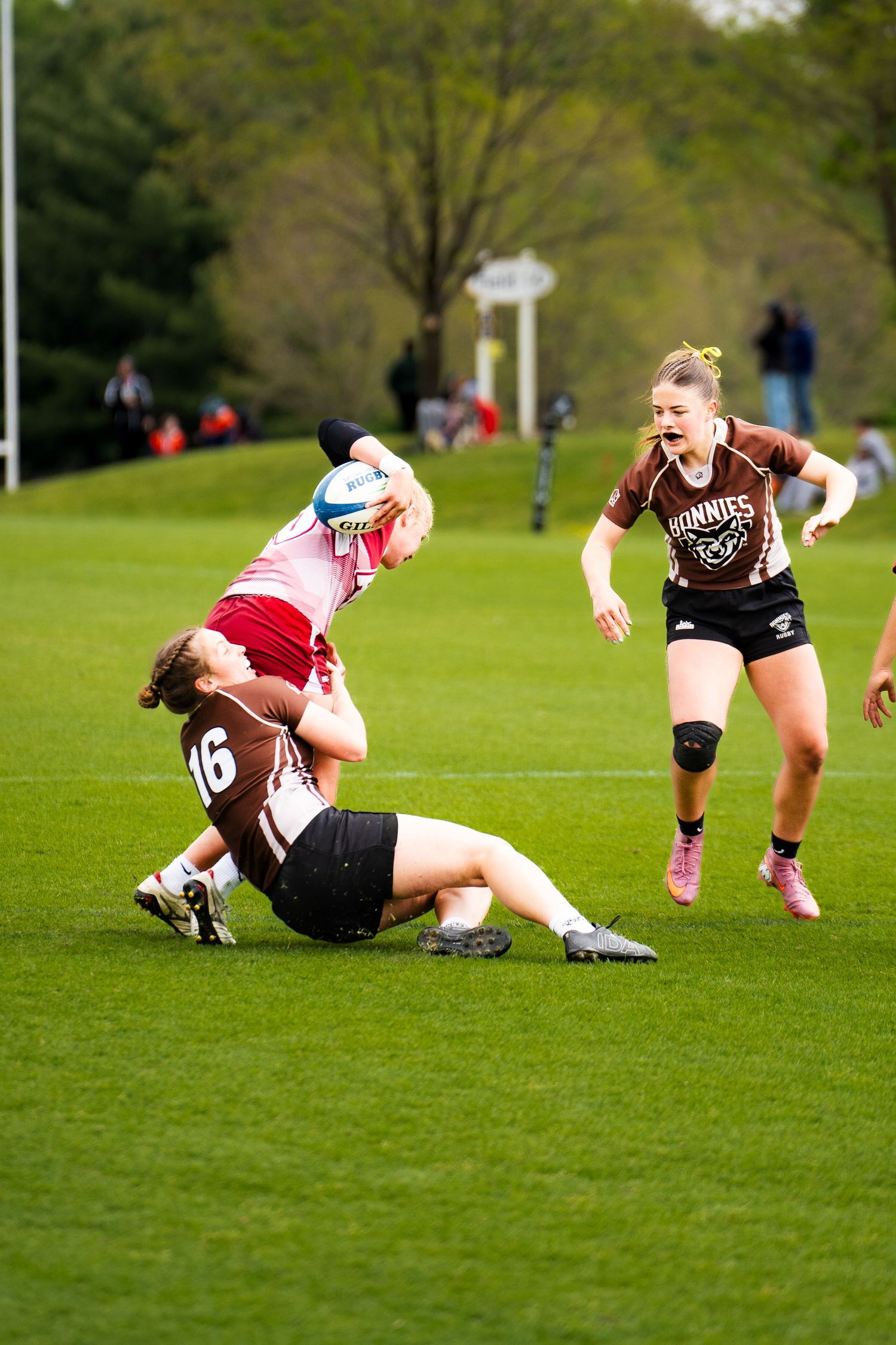 A rugby player in a brown jersey tackles a ball carrier in pink to the ground as a teammate runs in to help on a green field.