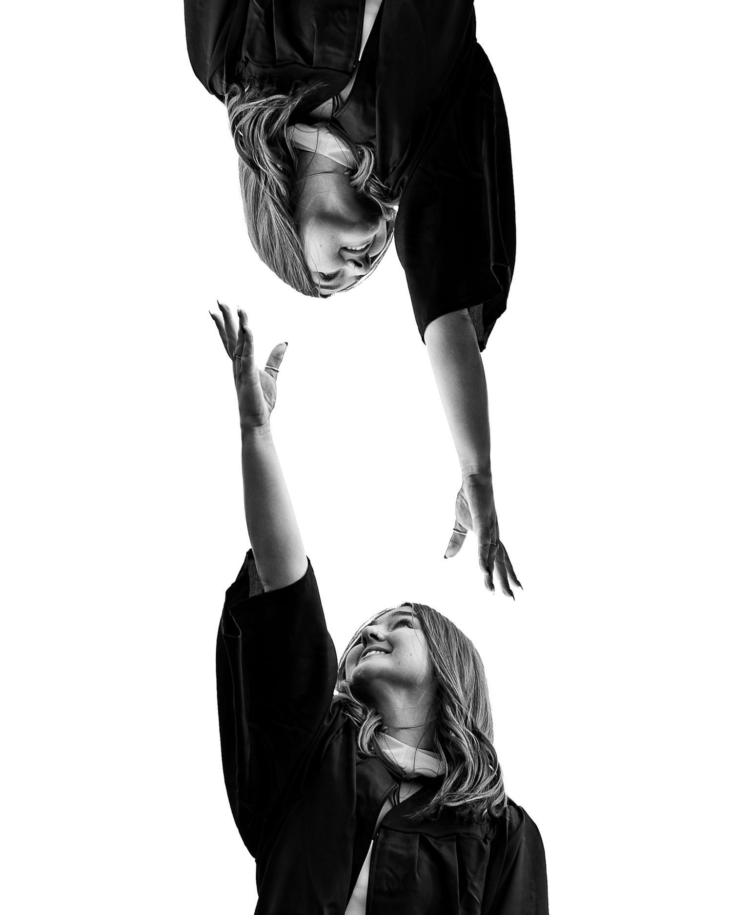 Black and white mirrored image of a woman in a graduation gown reaching upward, with her inverted reflection reaching down above her.