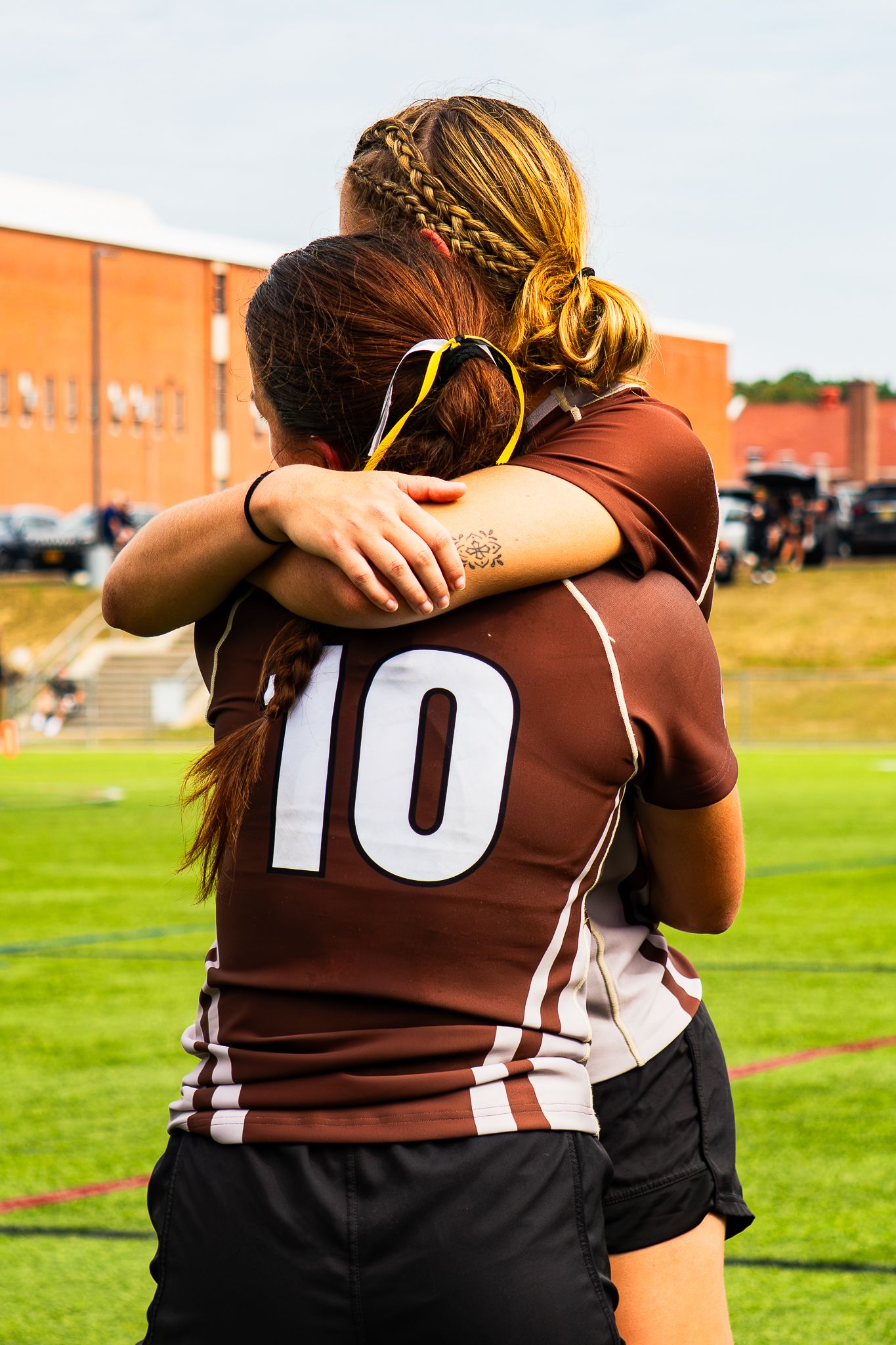 Two women rugby players in maroon jerseys embrace on a grassy field, one wearing number 10 with sunglasses on her head.