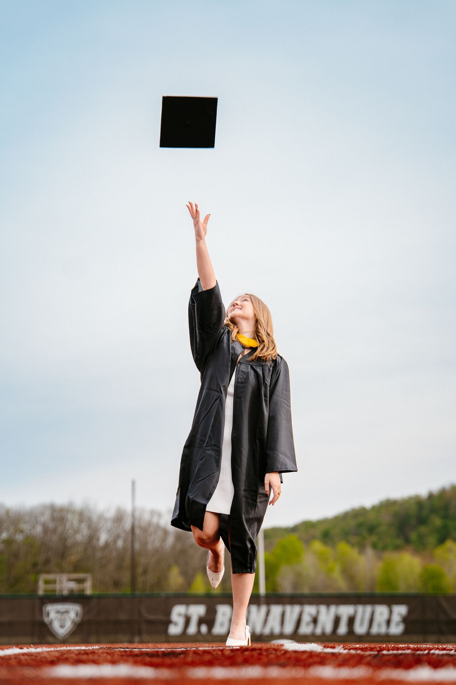 Graduate in a black gown tosses her cap high into the air on a baseball field at St. Bonaventure with hills in the background.