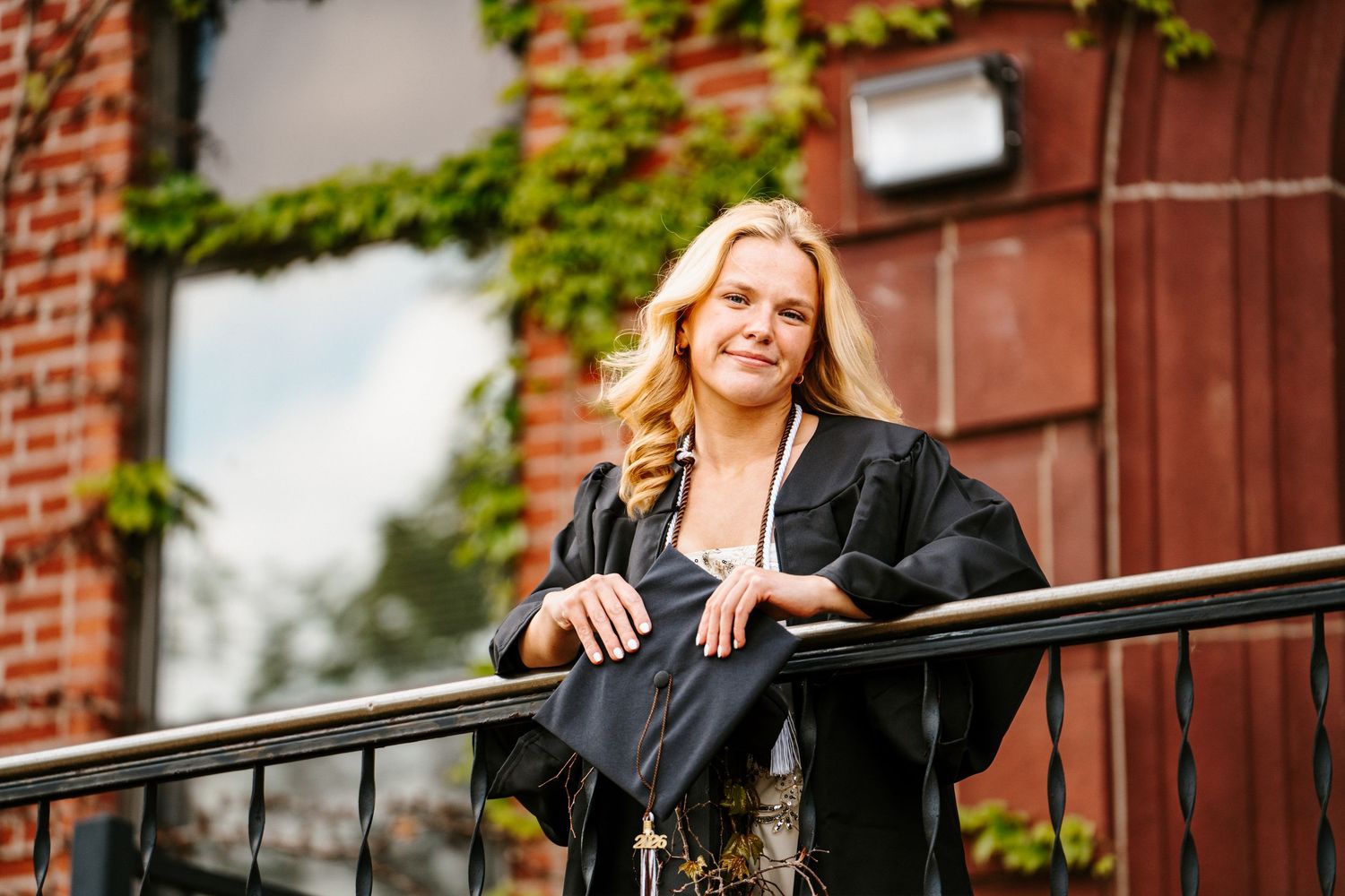 Blonde graduate in a black gown rests her arms on a railing and holds her cap in front of an ivy-covered brick wall.