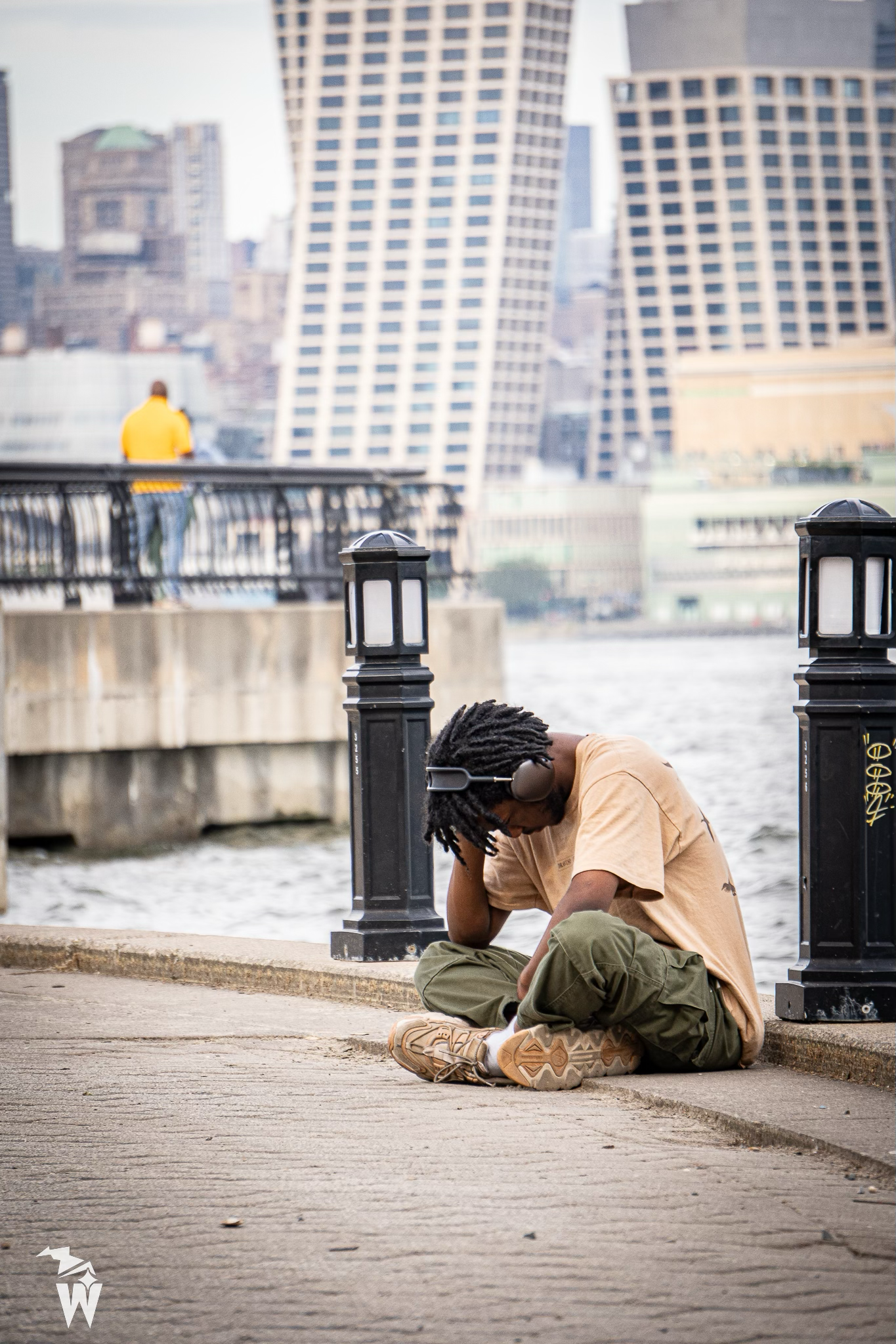 Young man in a tan shirt and olive pants sits on a waterfront walkway with his head down, with city buildings across the water.