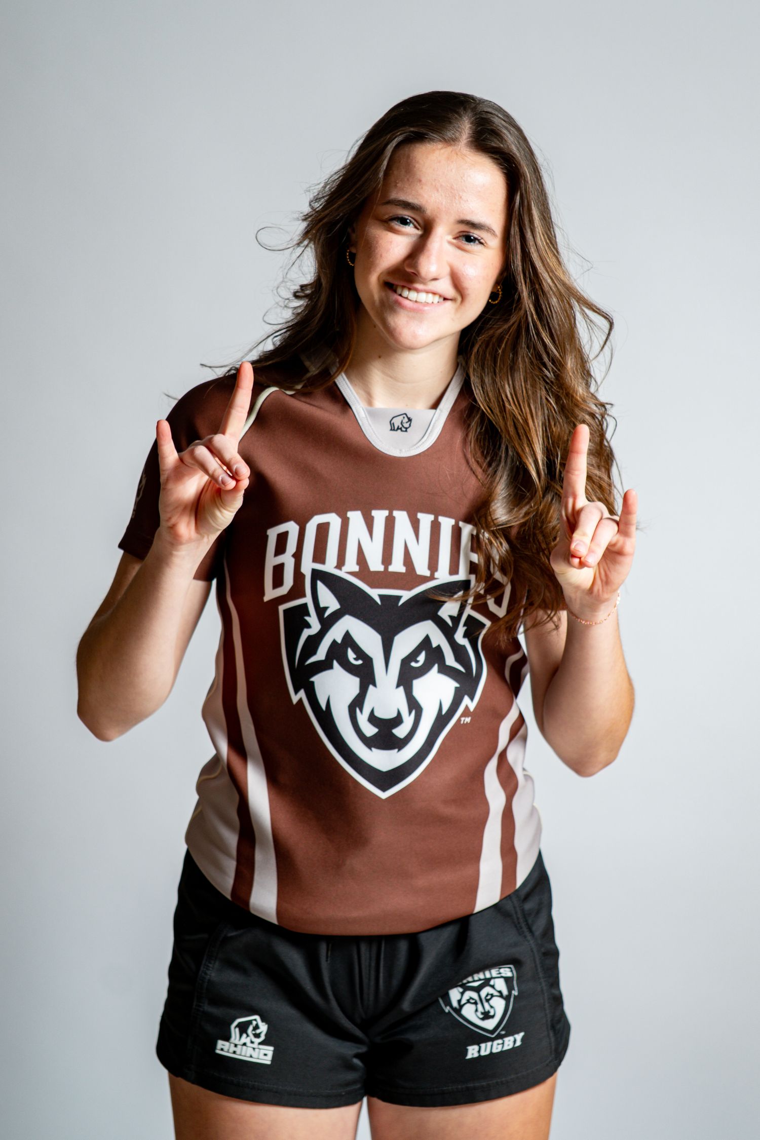 Smiling rugby player in a brown Bonnies wolf logo jersey and black shorts holds up two fingers on each hand against a white background.