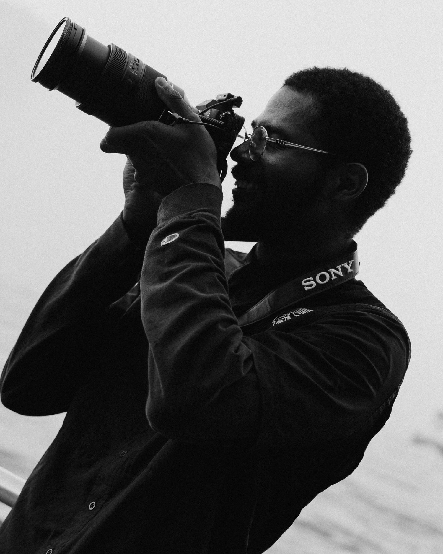 Black and white photo of a bearded man with glasses holding a Sony camera with a telephoto lens to his eye near the water.