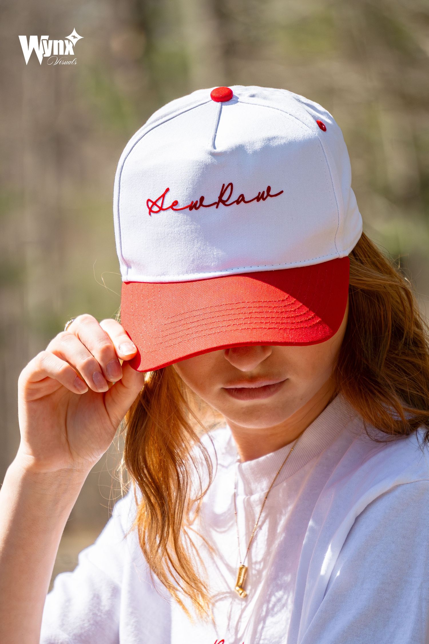 Woman tilts a white and red Sewlaw snapback cap with red script embroidery over her eyes while wearing a white tee and gold necklace.