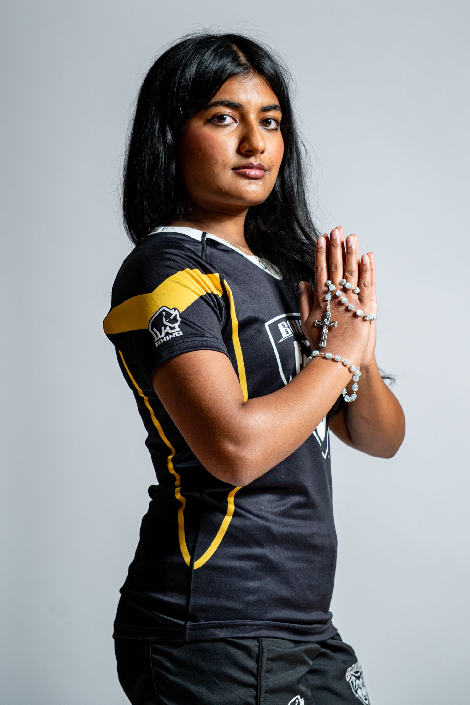 Young woman in a black and gold rugby jersey presses her hands together holding beaded bracelets against a plain gray backdrop.