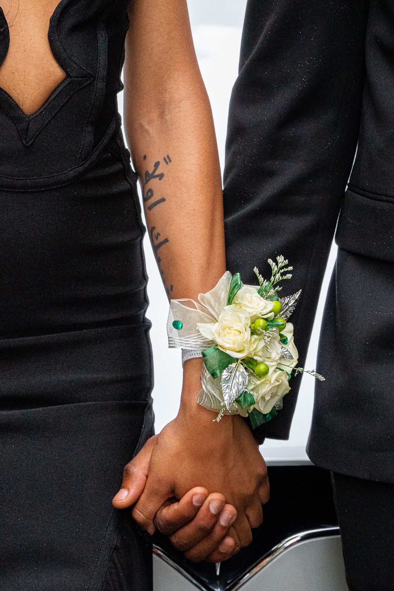Couple in black formal attire hold hands, the woman wearing a white rose wrist corsage with green accents and an arm tattoo.