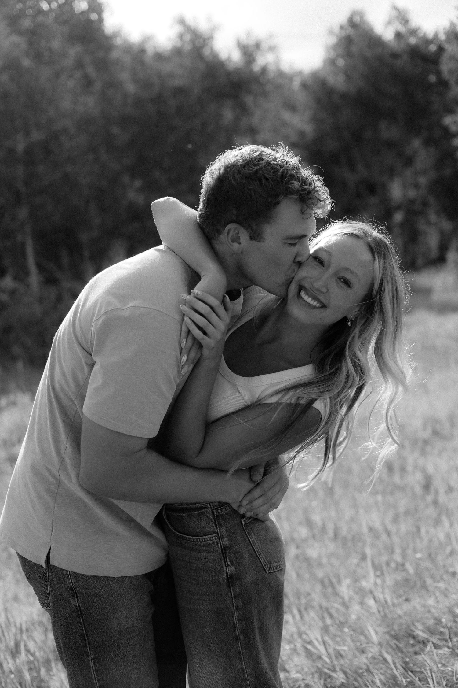 A black and white image showing a couple embracing in a field with trees in the background.