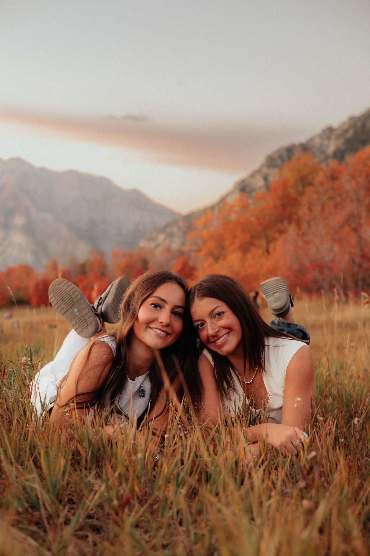 Two friends lay in autumn grass at sunset with mountains and fall foliage in the background.