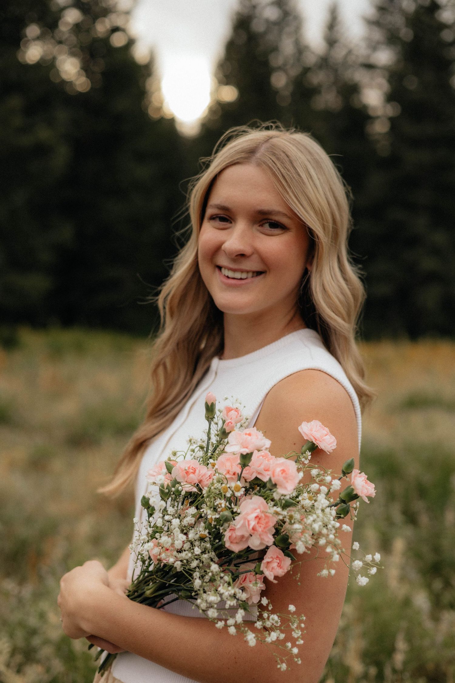 A smiling young woman with long blonde hair holds a bouquet of pink and white flowers in a field.