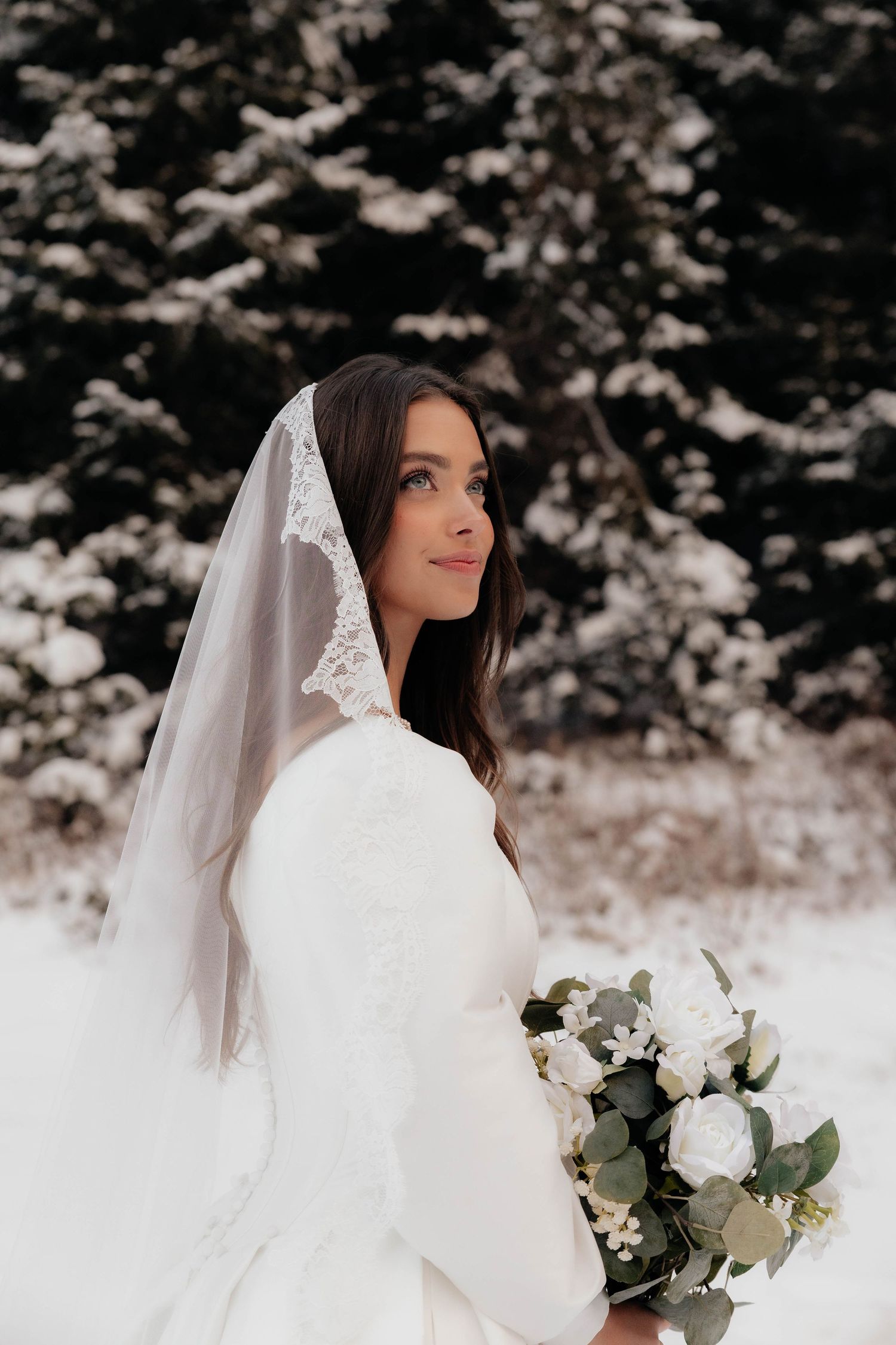 A bride in a white wedding dress and veil holds a bouquet of white flowers against a snowy winter backdrop.