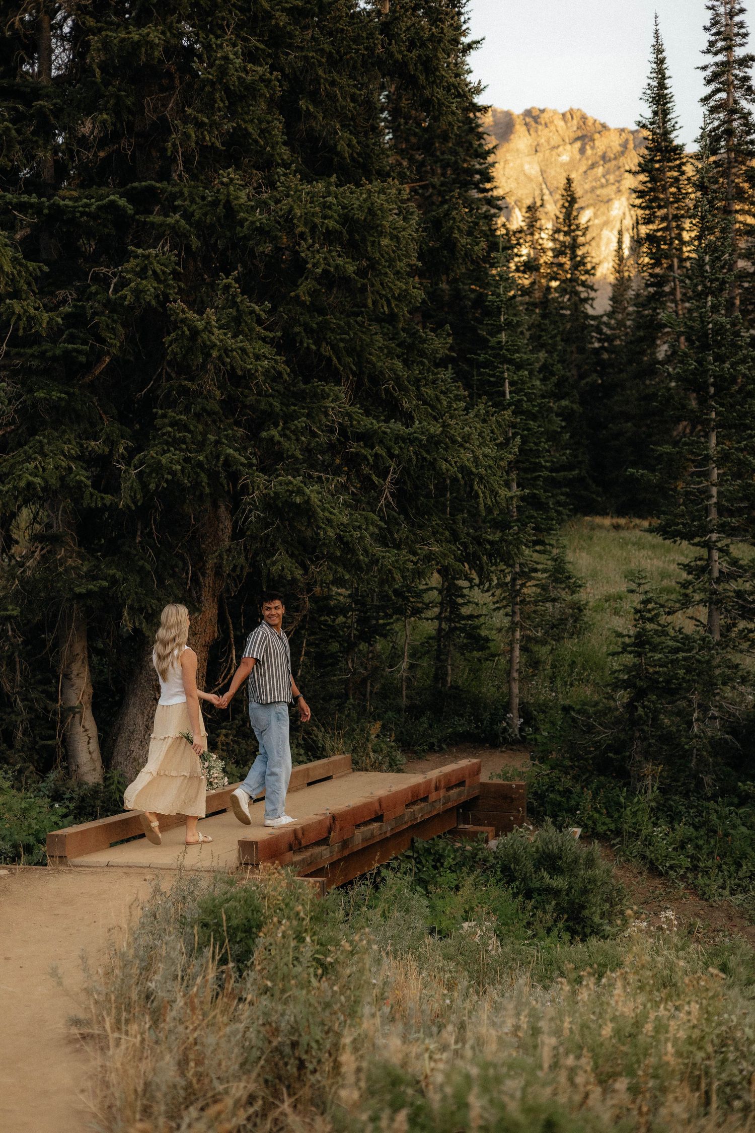 A couple sits on a fallen log surrounded by evergreen trees and mountain scenery at sunset.