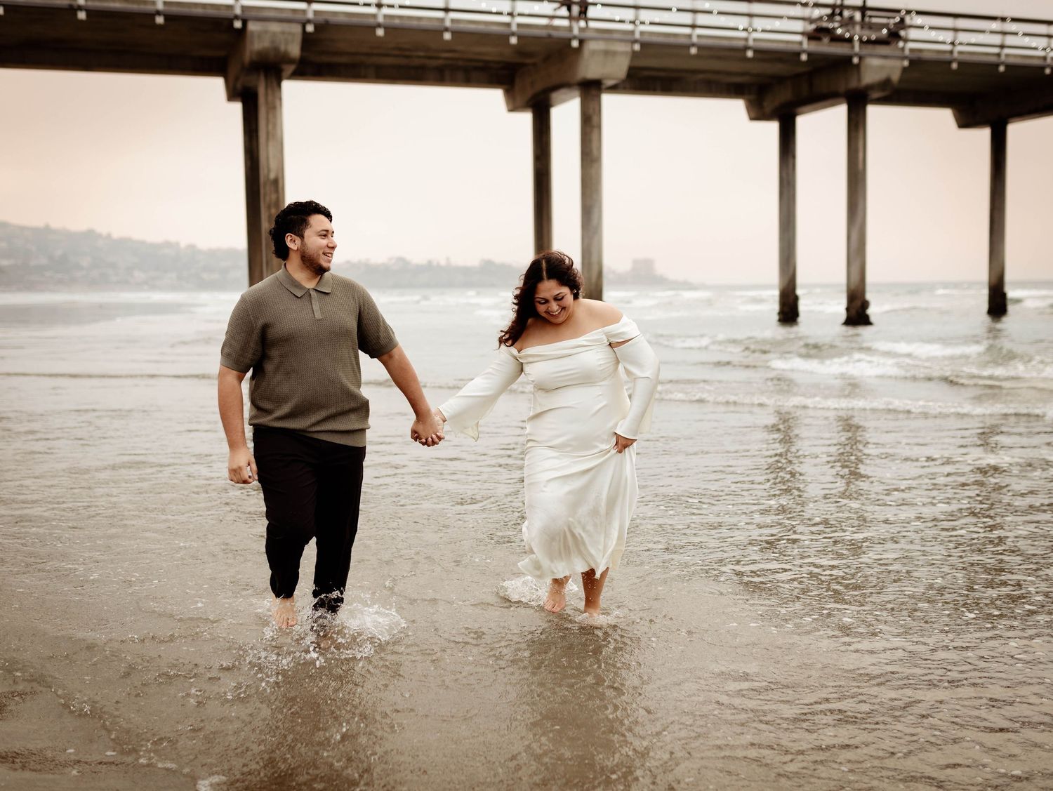 well-dressed engaged couple running along the shore in the water at the beach, holding hands and laughing together at Scripps Pier