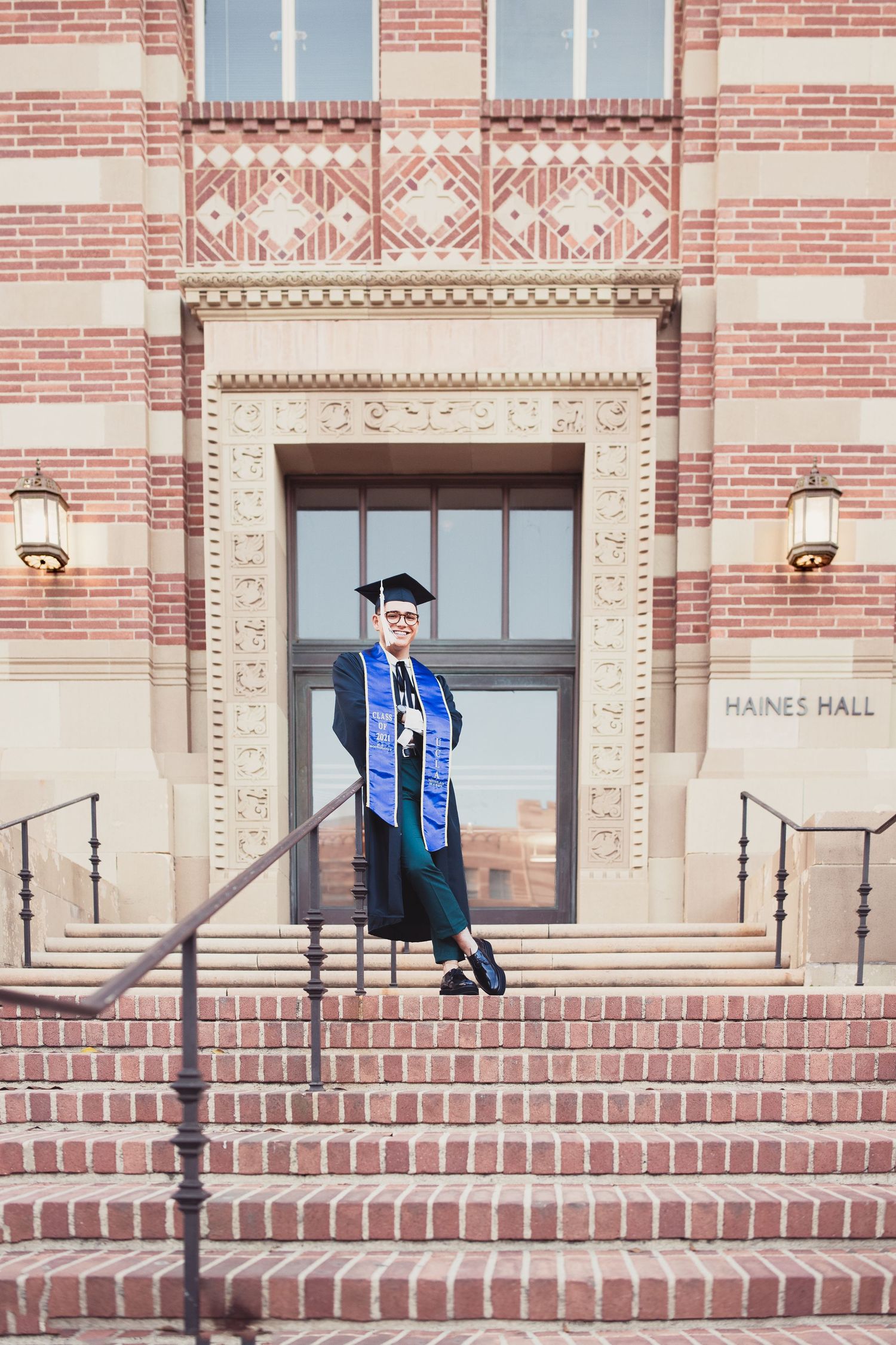 Graduate in cap and gown stands confidently on steps outside UCLA Haines Hall.