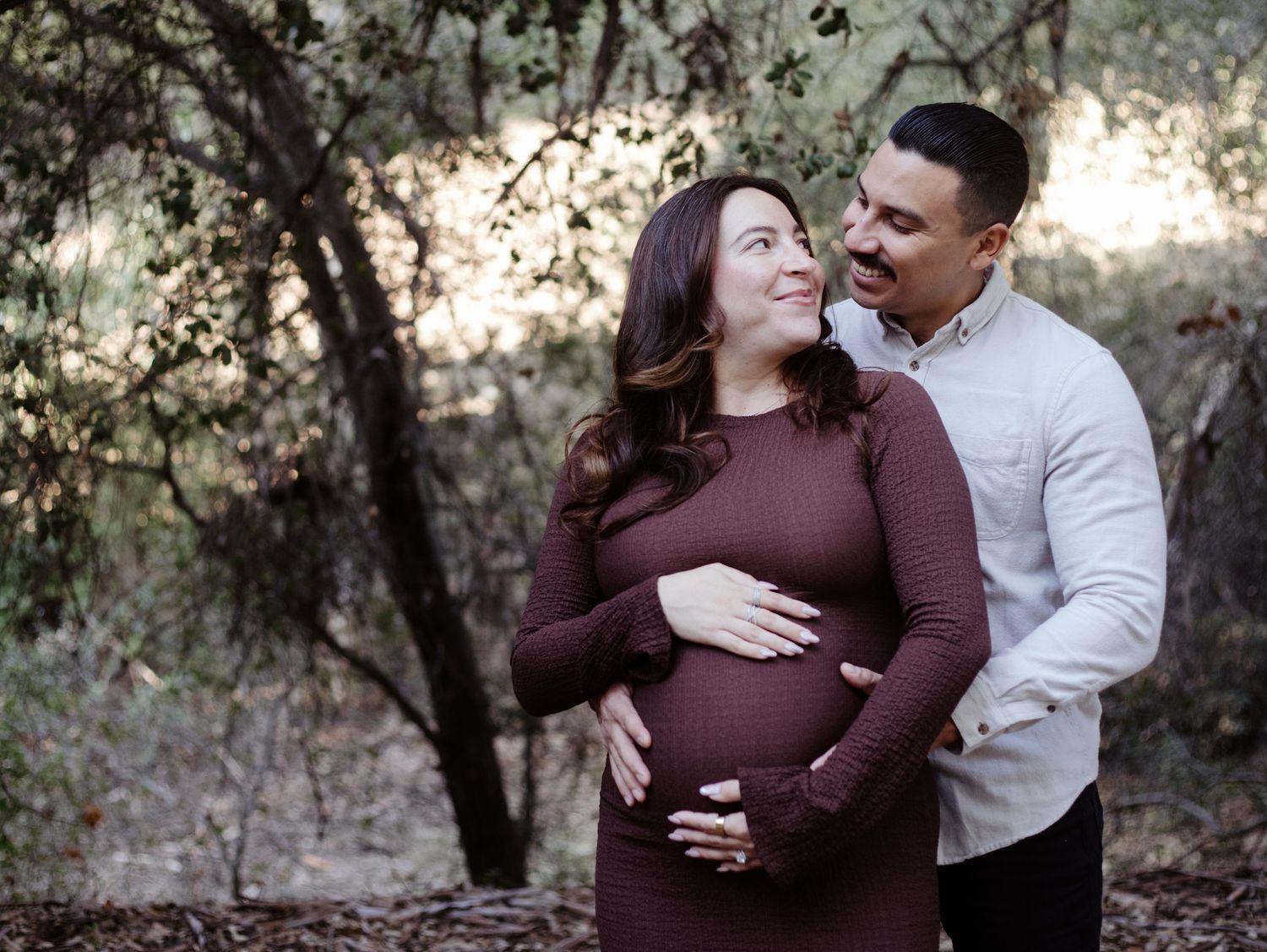 Pregnant couple standing together, smiling at each other during a rustic maternity session in the fall at Murphy Ranch Park in Los Angeles