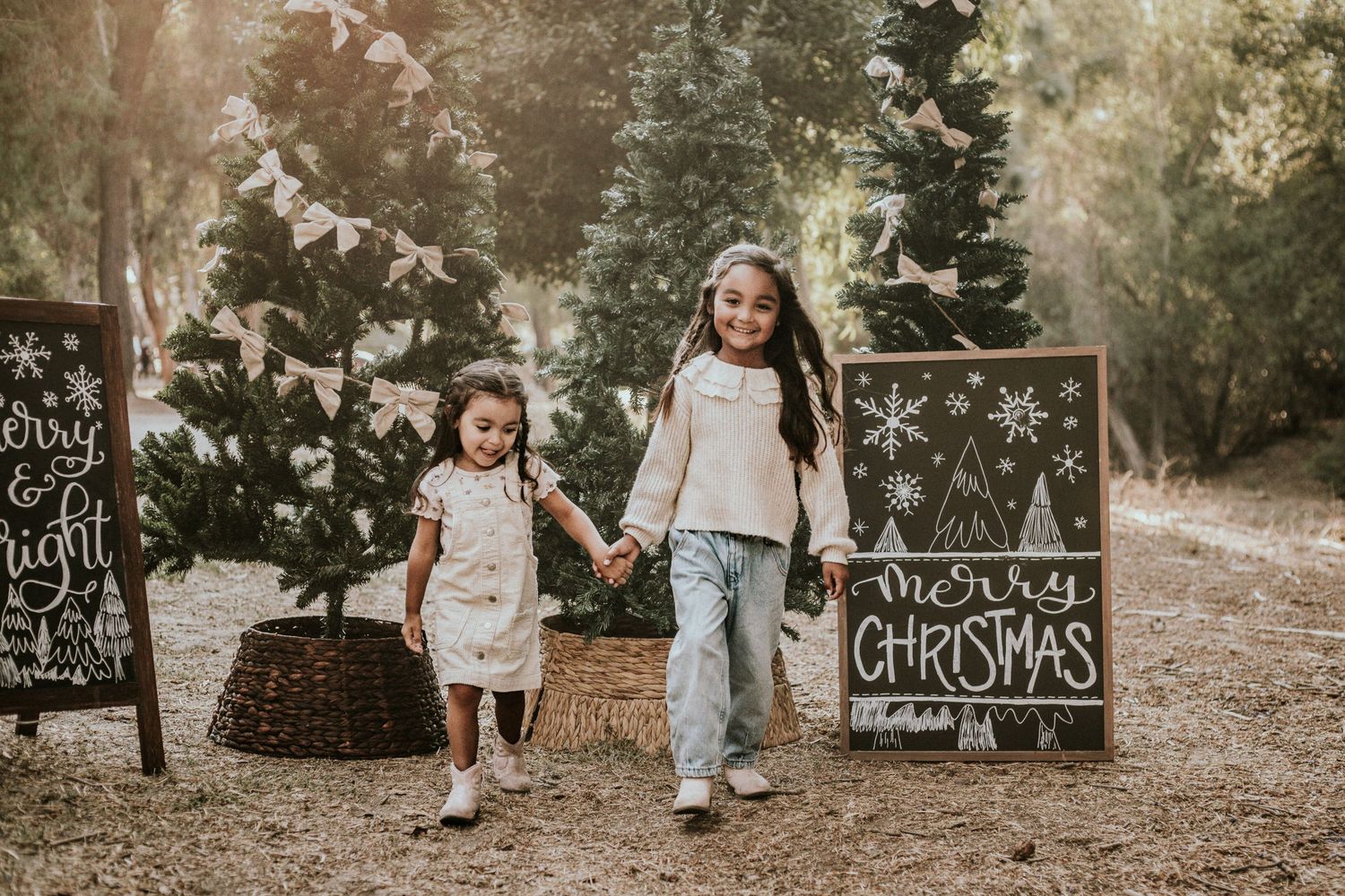 Two young girls holding hands walk past a Merry Christmas chalkboard sign among Christmas trees outdoors.