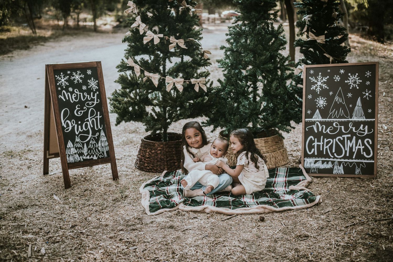 Three children sit on a plaid blanket between Christmas trees and chalkboard signs reading Merry & Bright and Merry Christmas.
