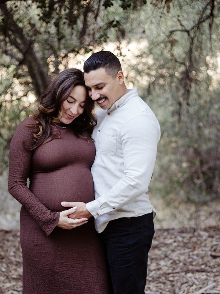 Man and pregnant woman stand in wooded area (Murphy Ranch Park) during a maternity session. They are smiling while looking at her baby bump
