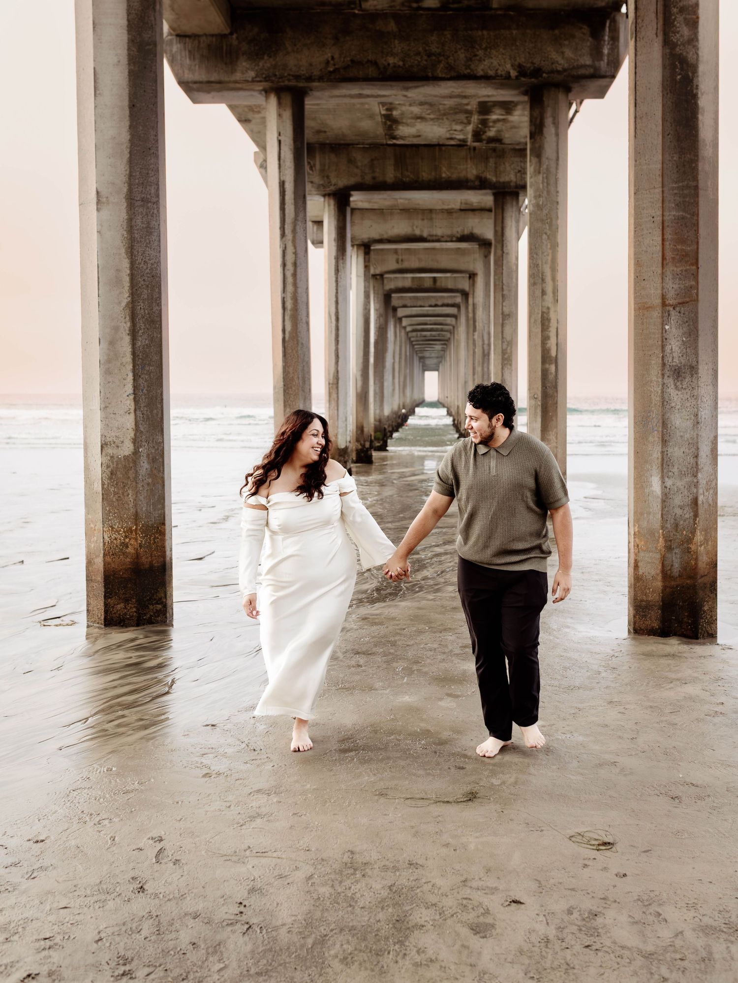 Scripps Pier San Diego Beach Engagement Portrait; couple is running under a pier at the beach; she is wearing an elegant, romantic dress
