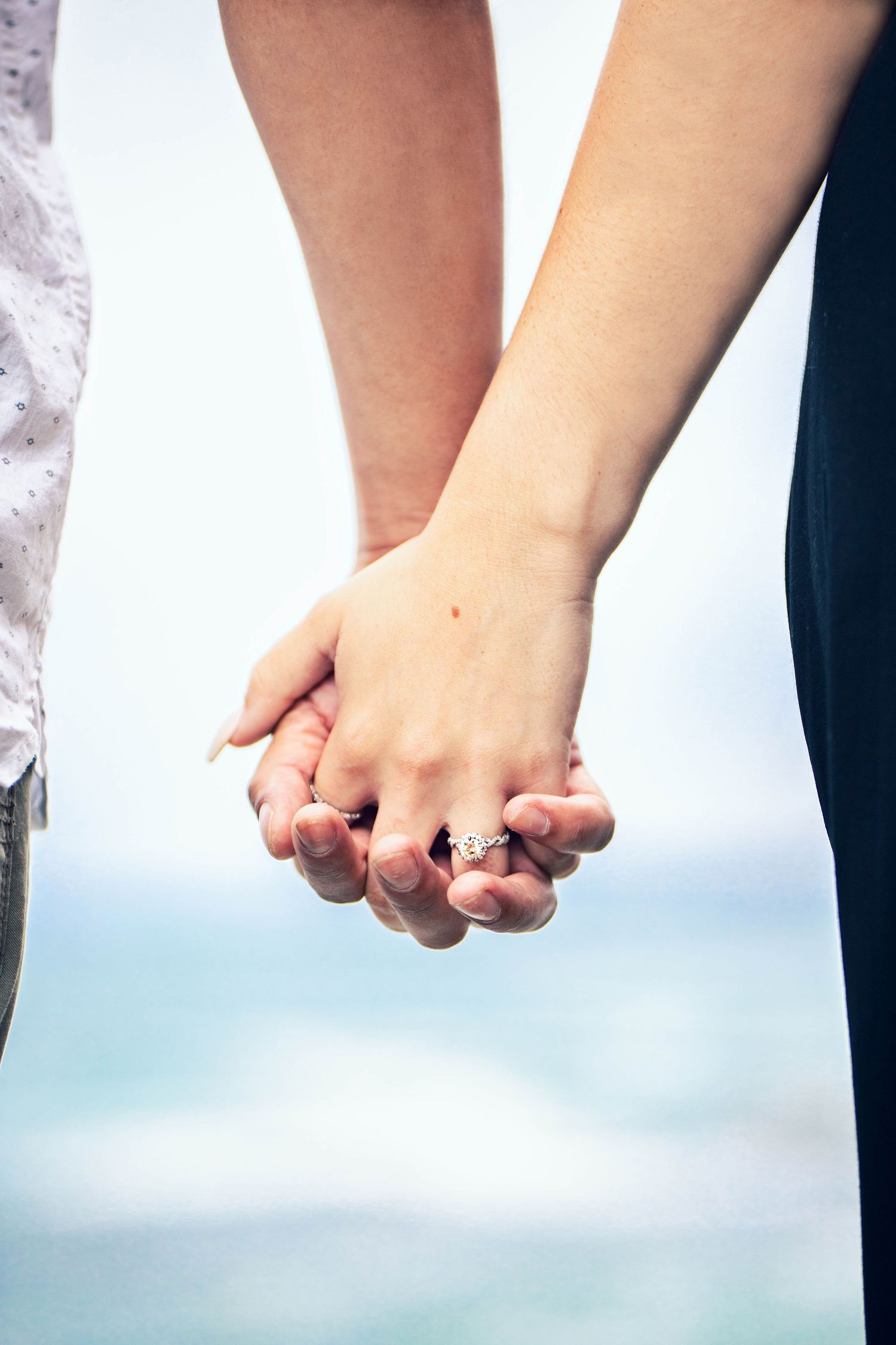 Close-up of an engaged couple holding hands, showcasing an engagement ring against a softly blurred ocean backdrop