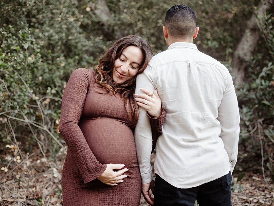 Maternity session; couple in fall clothes in a wooded area. His back to the camera, they lock arms. She smiles while holding her baby bump