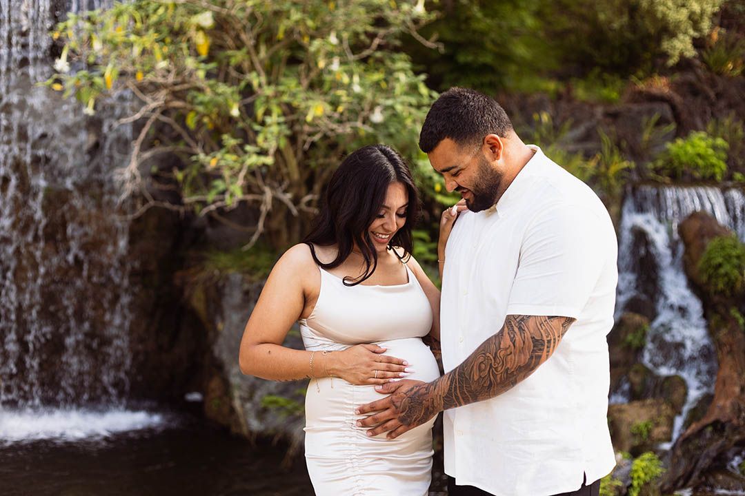 pregnant couple during maternity session at the Los Angeles Arboretum holding the baby bump as they each smile;  waterfall in background
