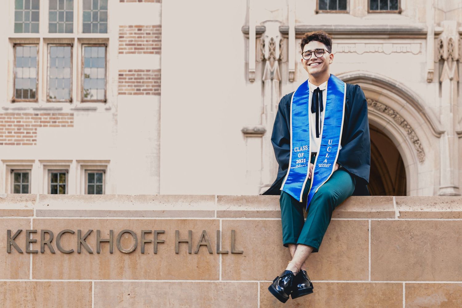 Smiling graduate in blue regalia sitting on Kerckhoff Hall wall at UCLA campus.