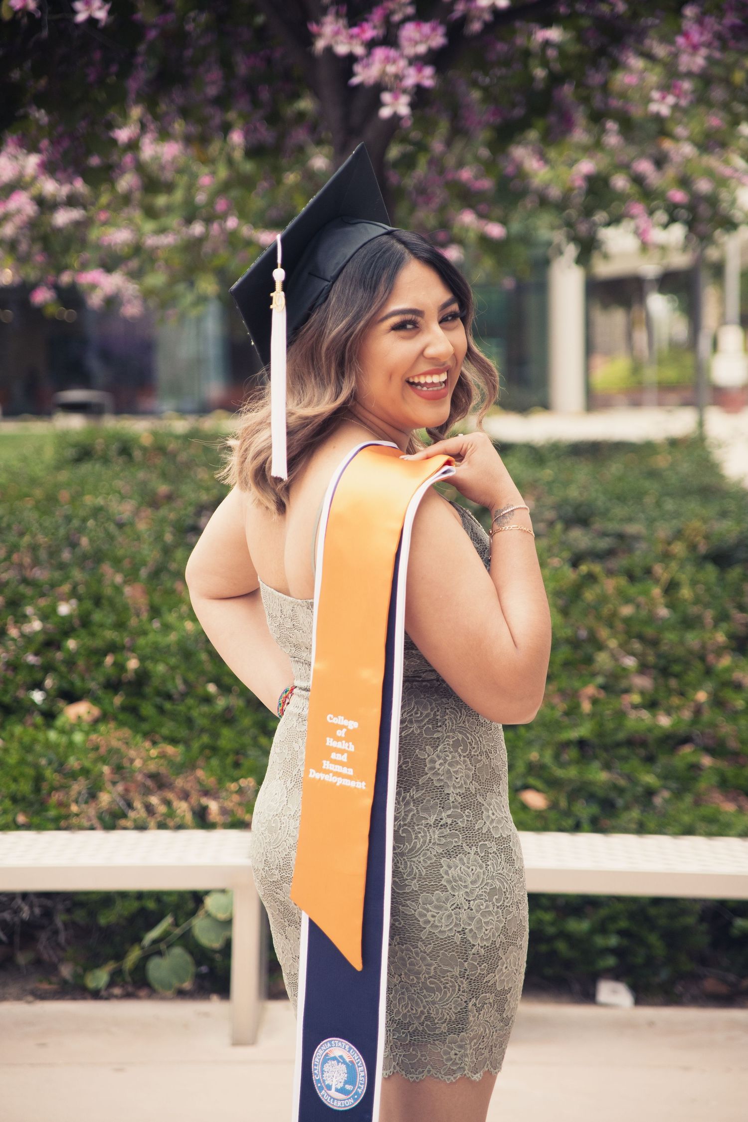 Smiling CSUF graduate wears cap and holds her sash with pretty greenery and florals in the background for this graduation session