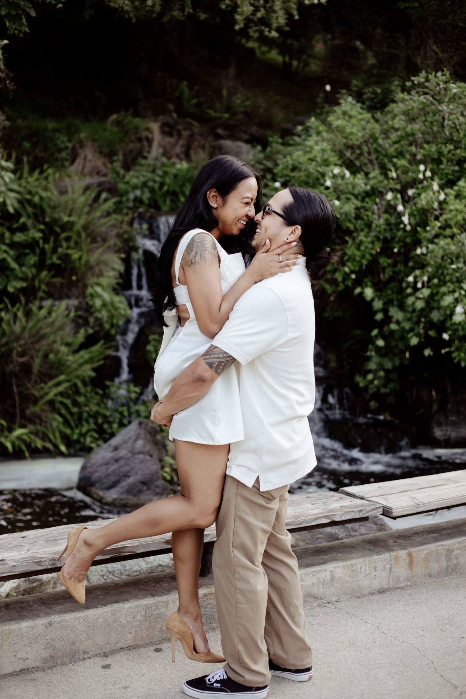 A well-dressed man is lifting a young woman up as they embrace and smile at each other. Lush greenery is in the background.