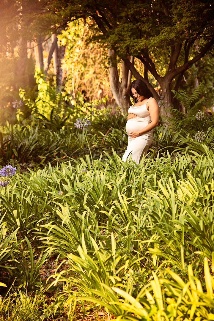 pregnant woman in cream dress standing in a green, grassy field at the Los Angeles Arboretum, looking at and holding her baby bump