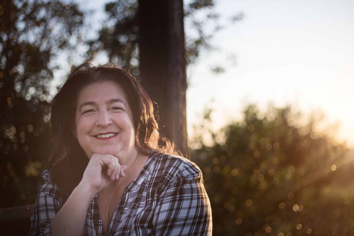 Woman is smiling at the camera as she sits in a wooded area during a sunset portrait session; the sunset glow is behind her.