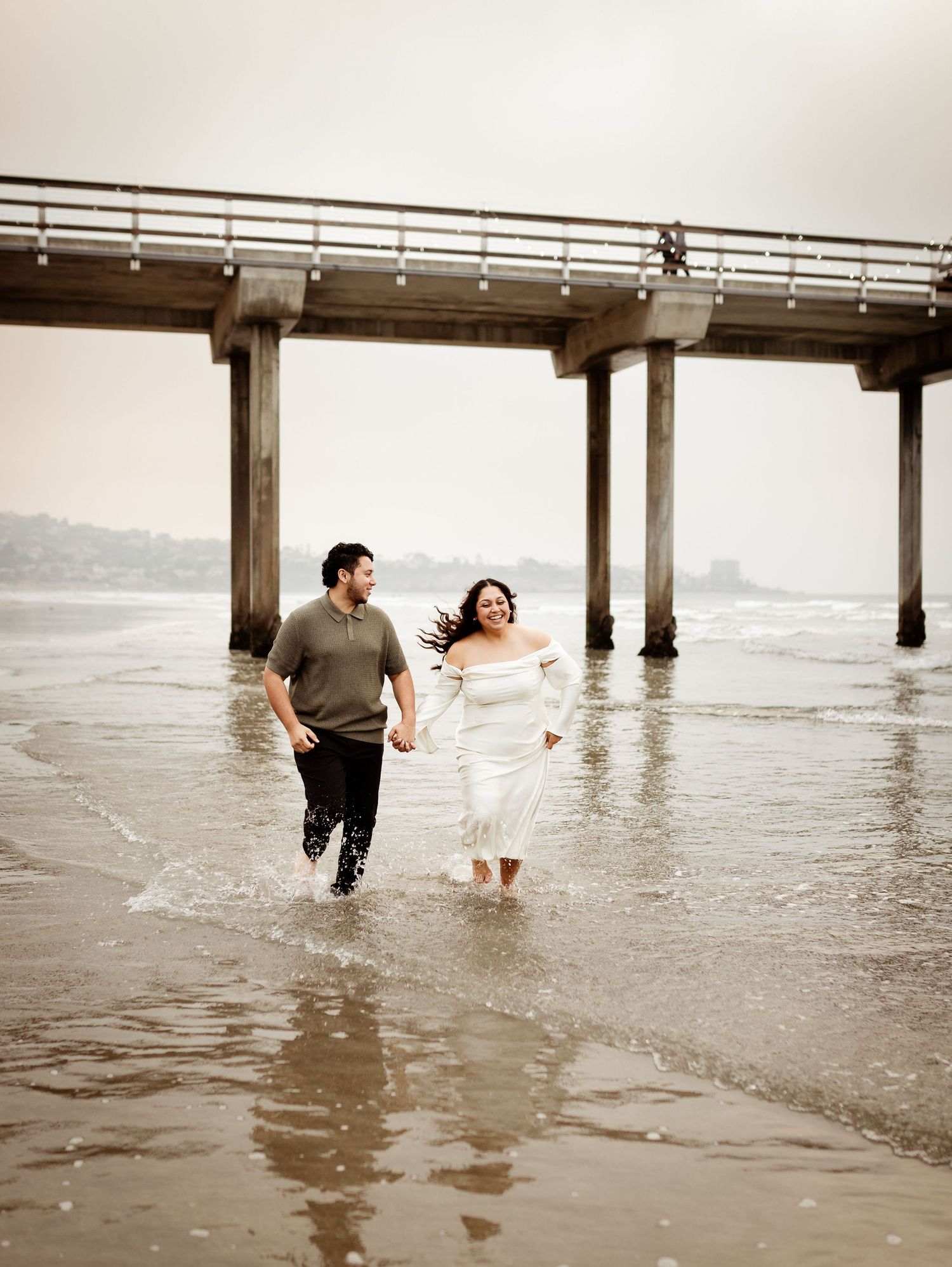 well-dressed engaged couple running along the shore in the water at the beach, holding hands and laughing together at Scripps Pier