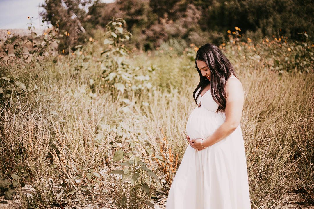 pregnant woman in a white, flowy dress standing in a wildflower field in Los Angeles, gazing poignantly at her baby bump while embracing it