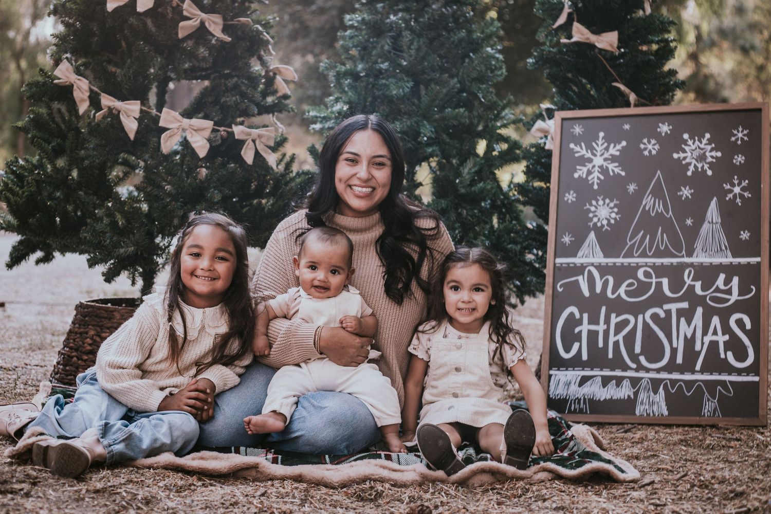 A mother poses with three young daughters in front of Christmas trees with a Merry Christmas chalkboard sign.
